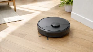 Robot vacuum with a raised LiDAR sensor turret cleaning a hardwood floor near a rug and potted plant.